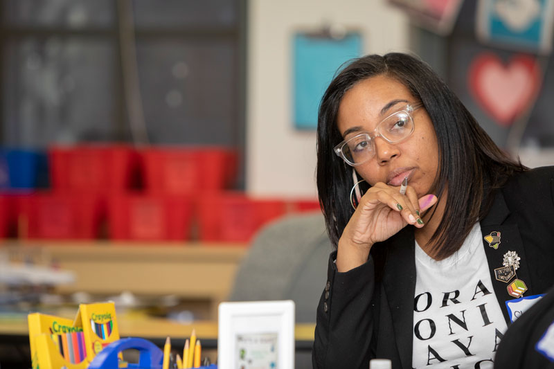 A teacher sitting at a table tilts her head to one side in thought. Around her is a colorful classroom.