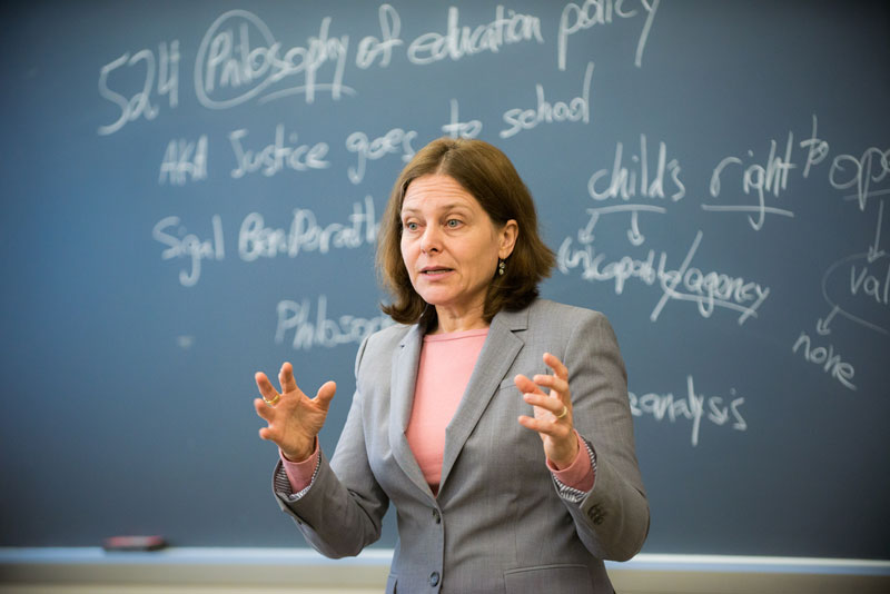 A professor addresses a class, standing in front of a blackboard with white chalk writings related to philosophy and education.