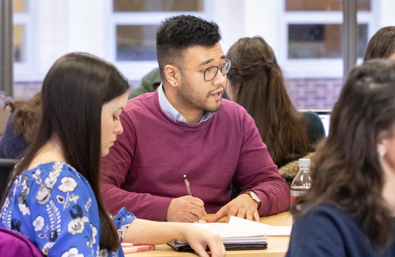 Two graduate students are sitting at a desk in a classroom, with other students seen in the background. One student takes notes while conversing, the other student looks down.