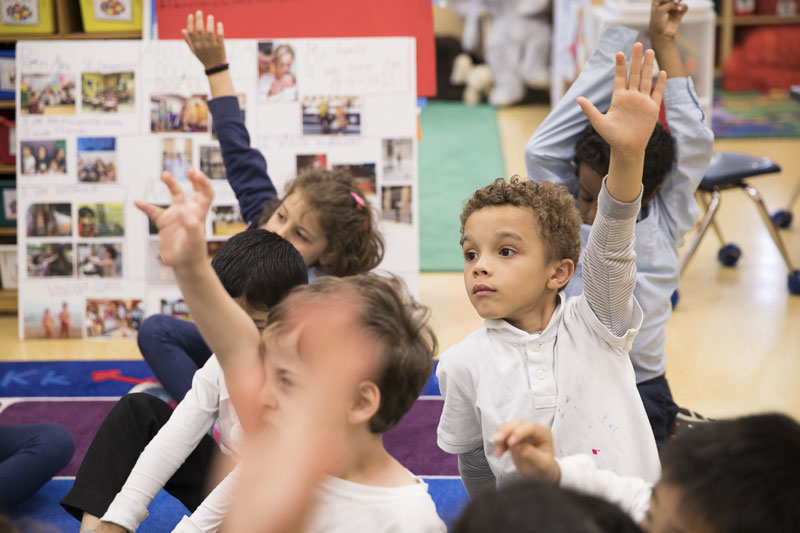 Elementary school children sit on the floor with their hands raised inside a colorful classroom.