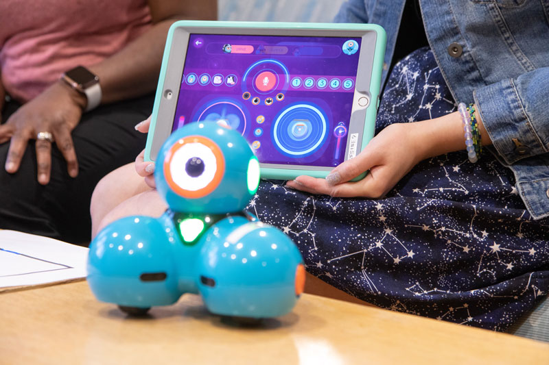 A blue robotic device with three spheres sits on a table. Behind it, an individual holds up a tablet device showing illuminated circles.