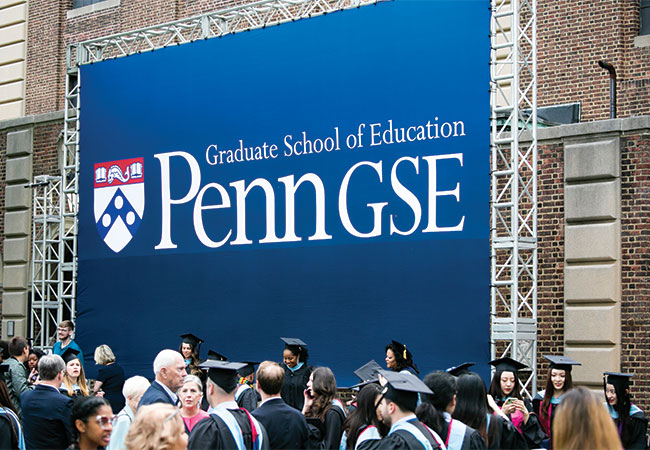 Penn GSE graduates in regalia and their family and friends gather in front of a large, blue sign with the Penn GSE logo.