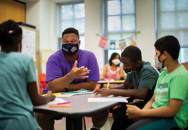 An instructor speaks with three K-12 students, seated with them around a table in a classroom as they write. All wear masks and colorful T-shirts.