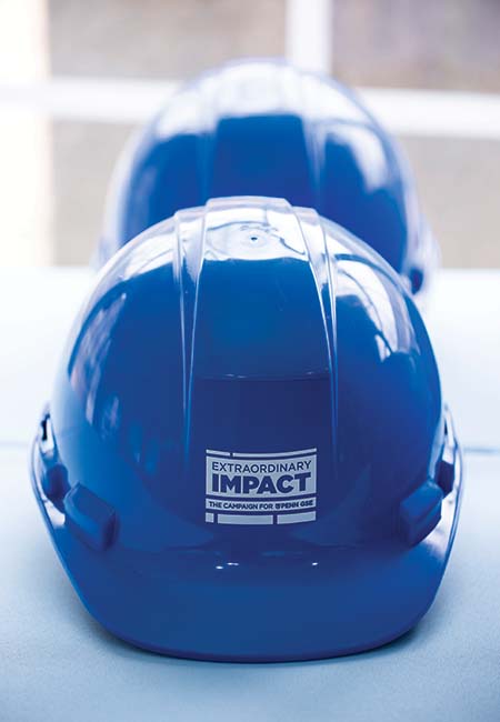 Two blue hard hats are placed on a flat surface. Text contained within the logo printed on the hard hat reads “Extraordinary Impact: The Campaign for Penn GSE.