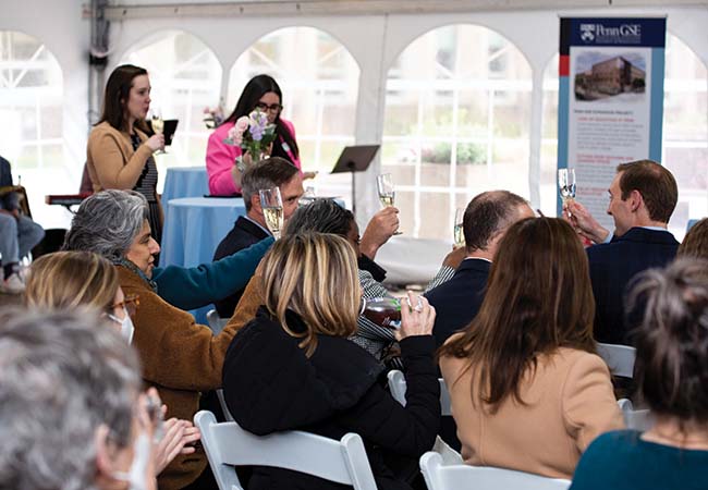 Audience members sit under a tent at the groundbreaking ceremony, some seated and some standing, raising their glasses for a toast.