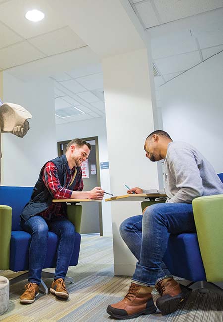 Two men wearing casual shirts and blue jeans are sitting across from each other in a student lounge area, smiling and looking at material on the desks in front of them.
